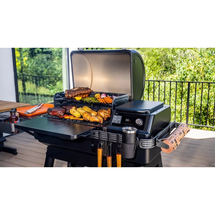 Traeger Ironwood wood pellet grill in a backyard setting with cooking lid open and grilling accessories visible.