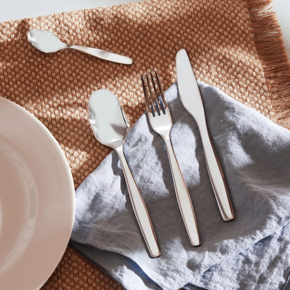 Alessi Itsumo 24-piece cutlery set with stainless steel forks, knives, spoons, and teaspoons arranged on a wooden table.