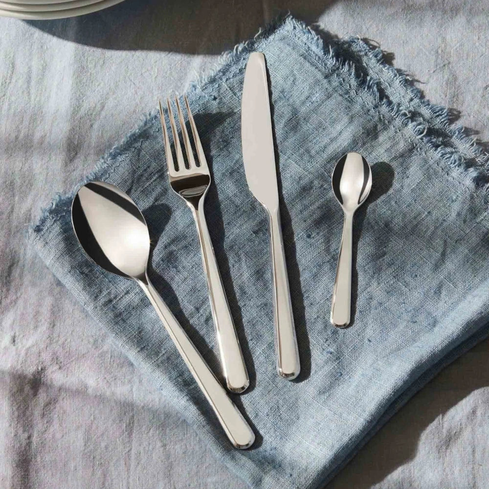 Alessi Amici 24-piece cutlery set with stainless steel forks, knives, and spoons arranged on a wooden surface.